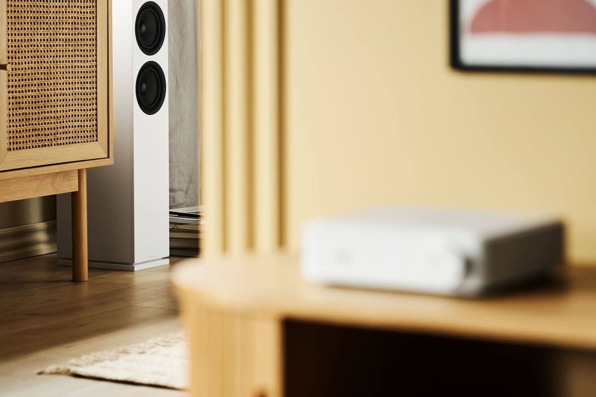 Diffusore da pavimento bianco accanto a un mobile in rattan, con un amplificatore sfocato in primo piano