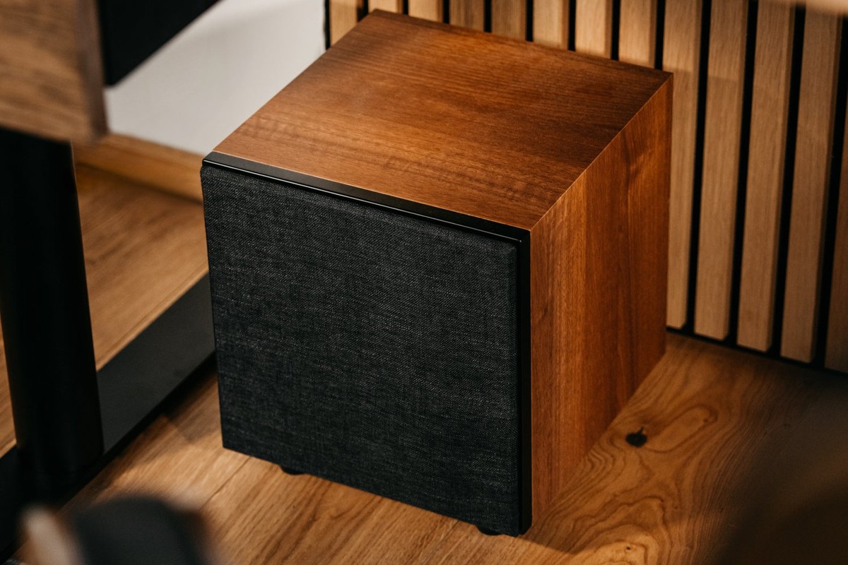 Walnut-finish subwoofer with fabric grille on a wooden floor next to a cabinet