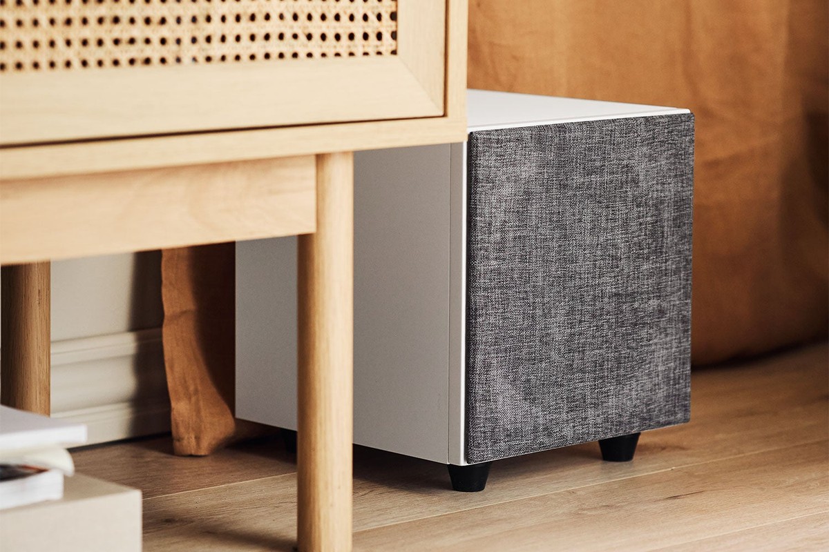 White subwoofer with grey grille tucked under a light wood console
