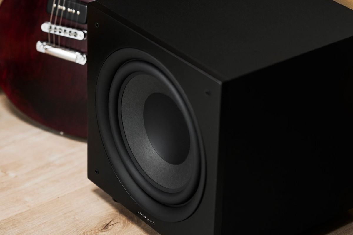 Caisson de basses noir posé sur un parquet, à côté d'une guitare électrique