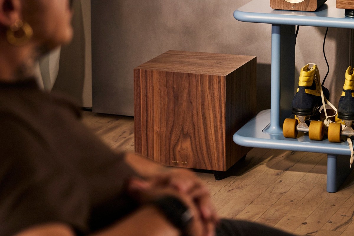 Subwoofer in Nussbaumfurnier auf dem Boden neben einem Regal im Wohnzimmer