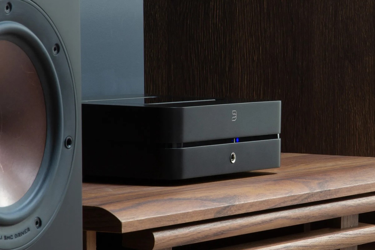 Black amplifier on a wooden cabinet in front of a floorstanding speaker
