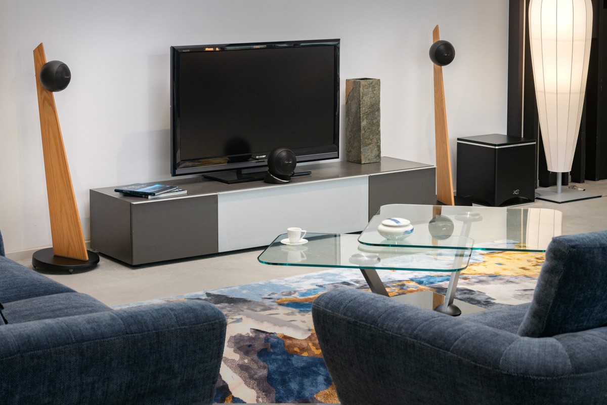 Soggiorno con televisore su mobile grigio, due satelliti su piedistalli in legno chiaro e subwoofer nero a terra sulla destra