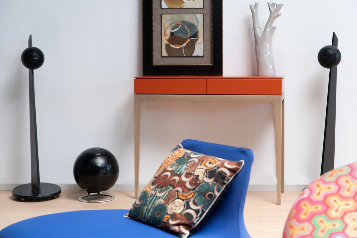 Black subwoofer between two iO3 speakers on stands, blue armchair in the foreground