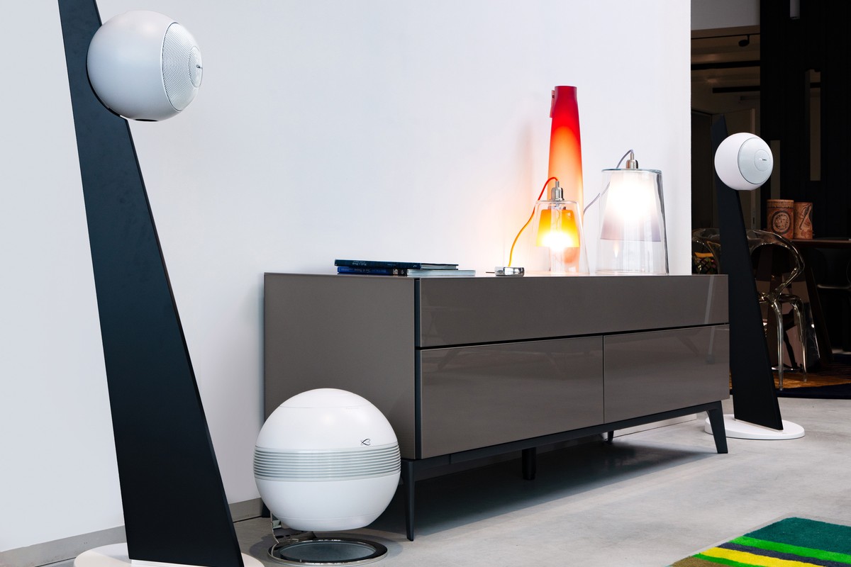 White subwoofer at the foot of a sideboard, flanked by two Riga 2 speakers on black stands