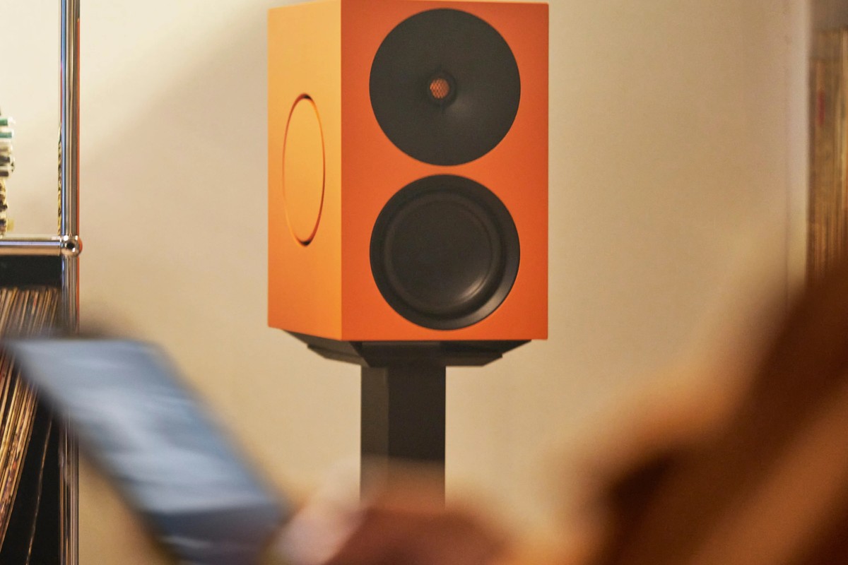 Oranger L/R X Lautsprecher auf einem Ständer, im Vordergrund ein unscharfes Smartphone