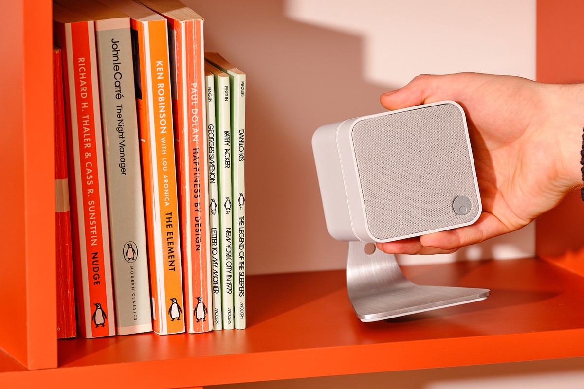 White speaker held in a hand above a stand, on a shelf next to books