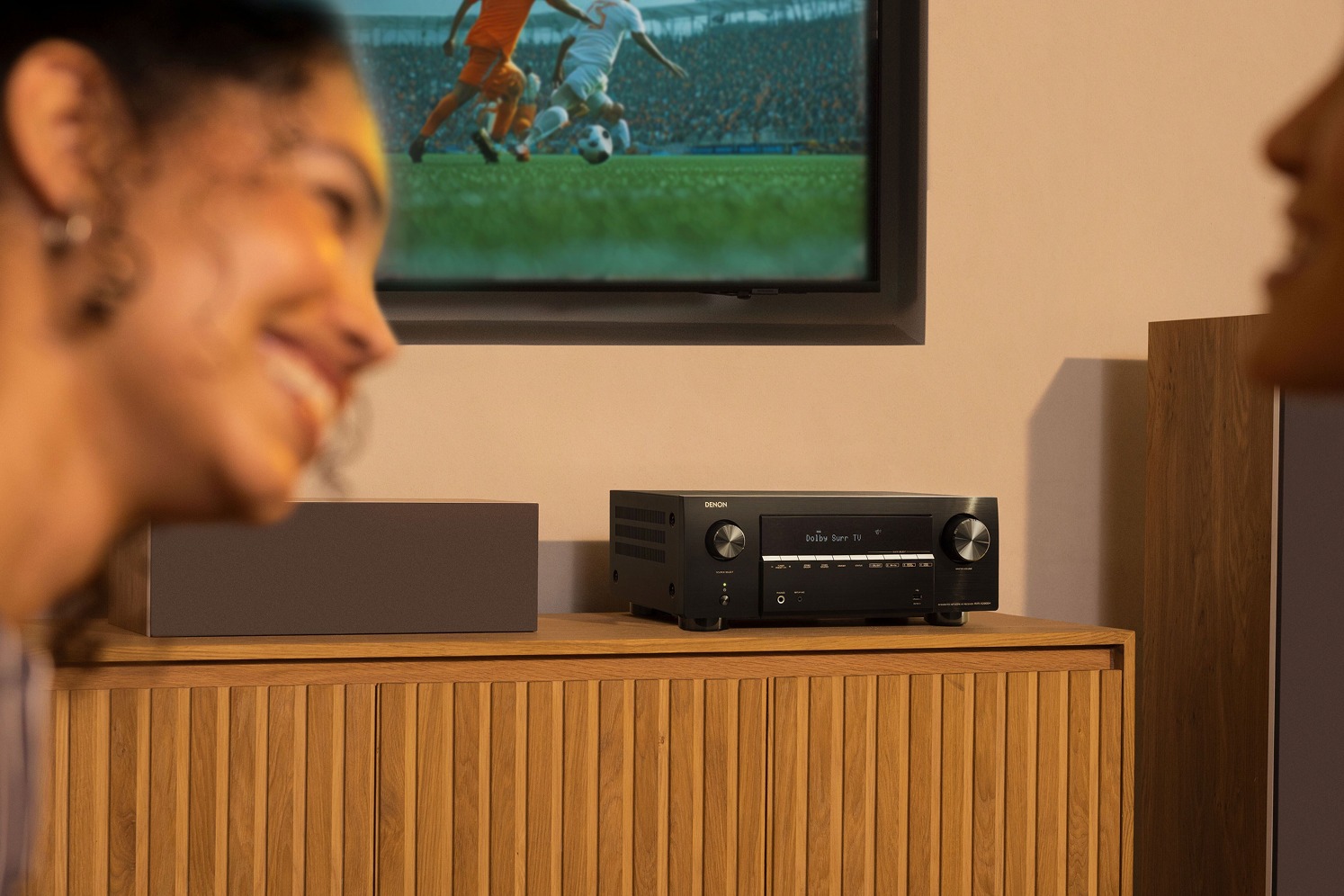 Spectatrice souriante devant un match, ampli-tuner et enceinte centrale sur le meuble
