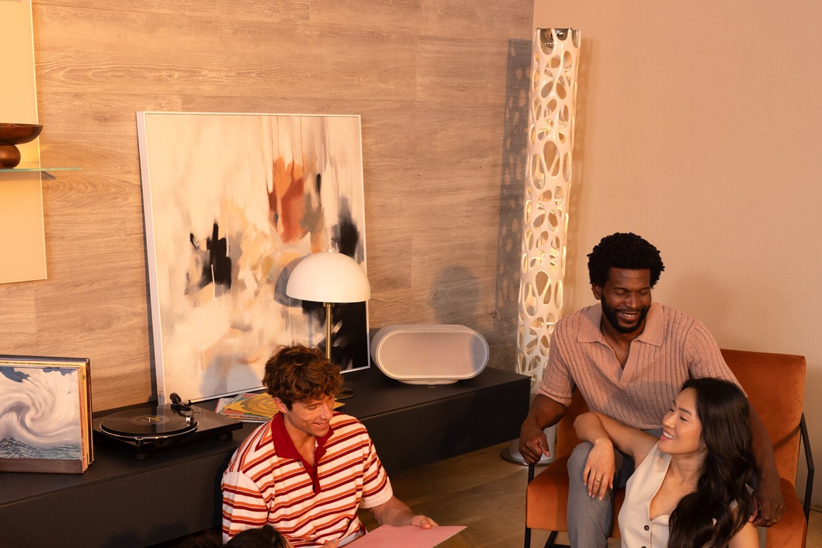Enceinte connectée beige clair sur un meuble bas près d'une platine vinyle, amis réunis au salon