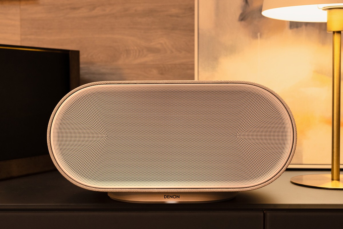 Enceinte beige clair vue de face sur un meuble sombre, à côté d'une lampe dorée