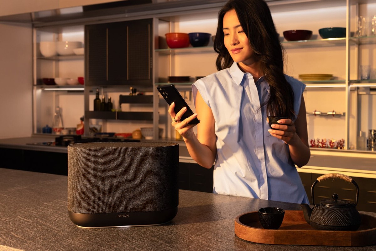 Enceinte connectée gris anthracite sur un plan de travail de cuisine, femme tenant un smartphone