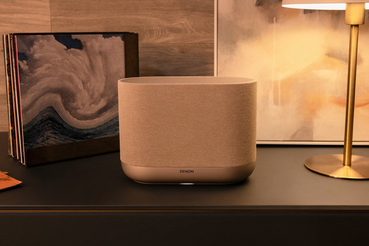 Enceinte sans fil beige clair sur un buffet sombre, à côté de vinyles et d'une lampe dorée