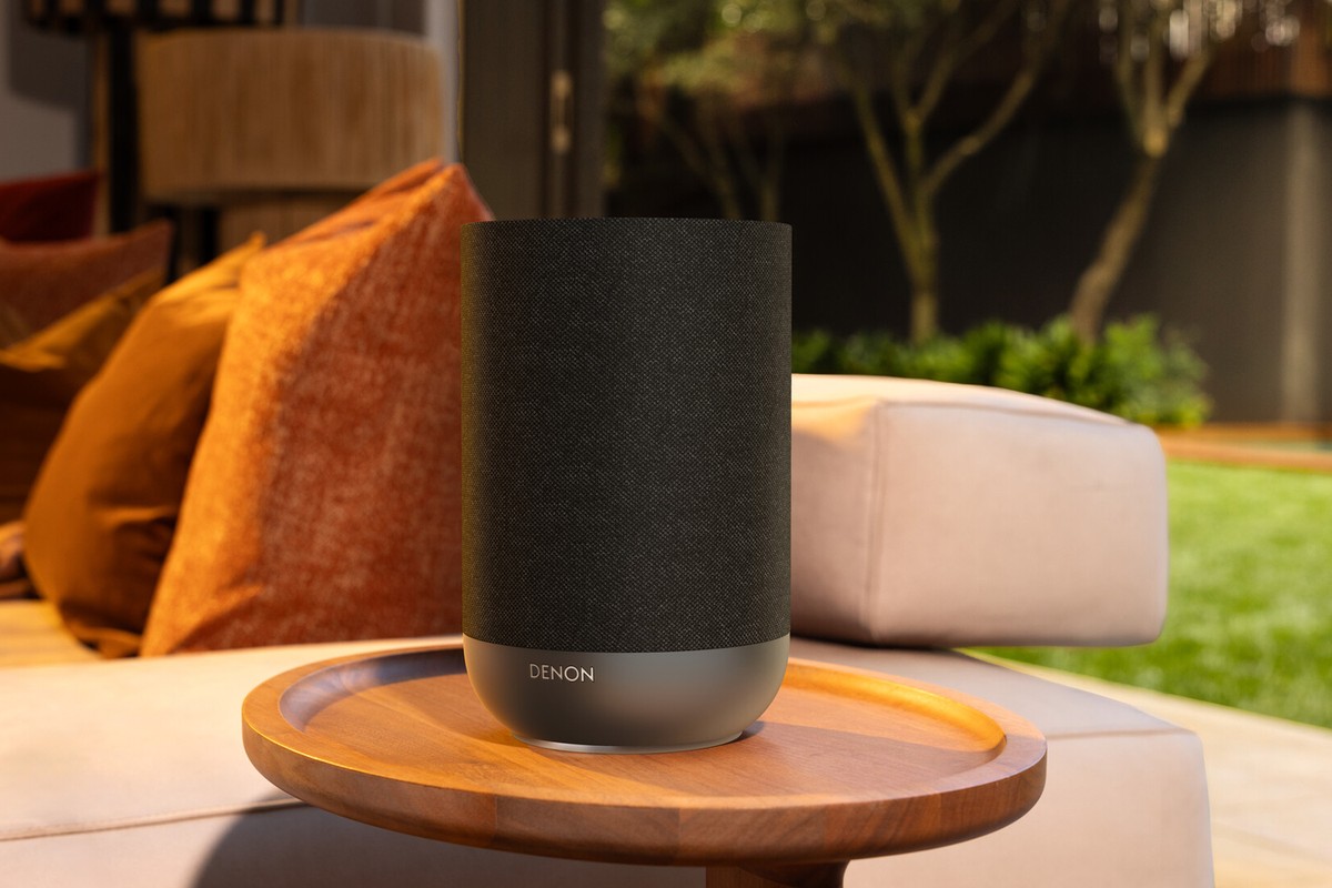 Kompakter anthrazitgrauer Wi-Fi-Lautsprecher auf einem Holztablett, Garten im Hintergrund