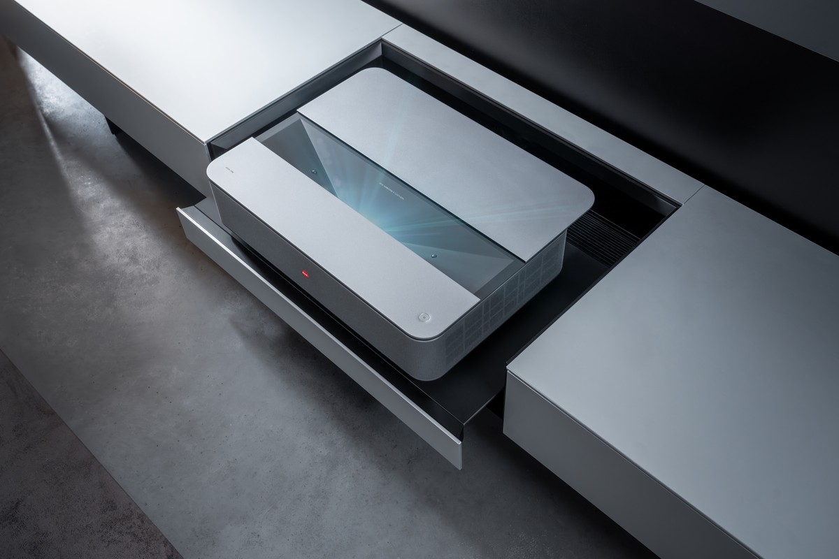 High-angle view of the projector with its lid open in a grey cabinet, lens visible