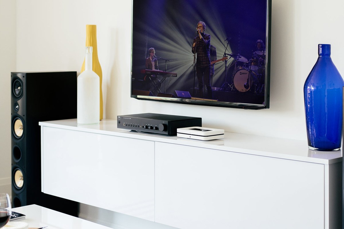NAD C 316BEE V2 amplifier on a white cabinet below a TV, between two floorstanding speakers