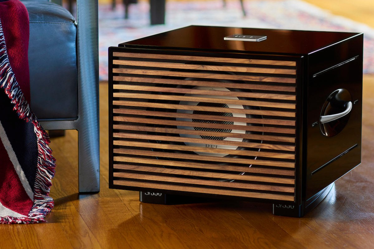 Black subwoofer with wood slats on a parquet floor, next to an armchair