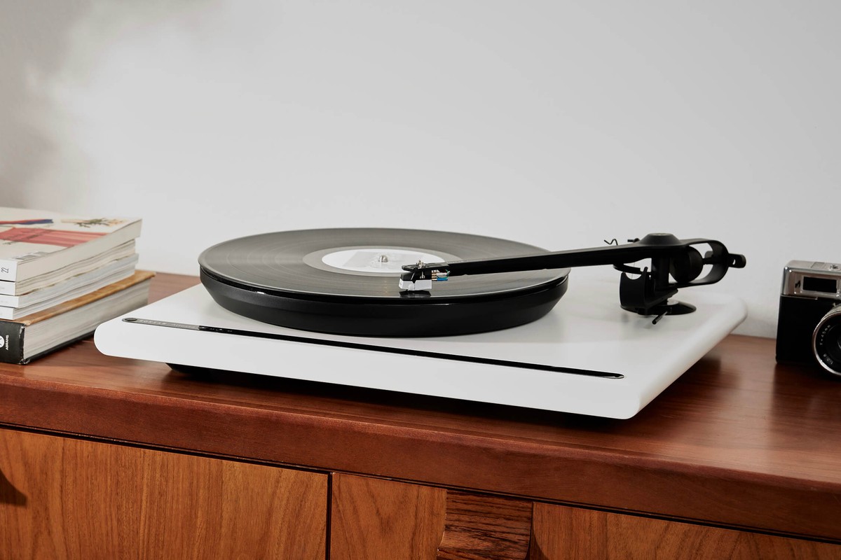 White turntable on a walnut sideboard, between a stack of magazines and a camera