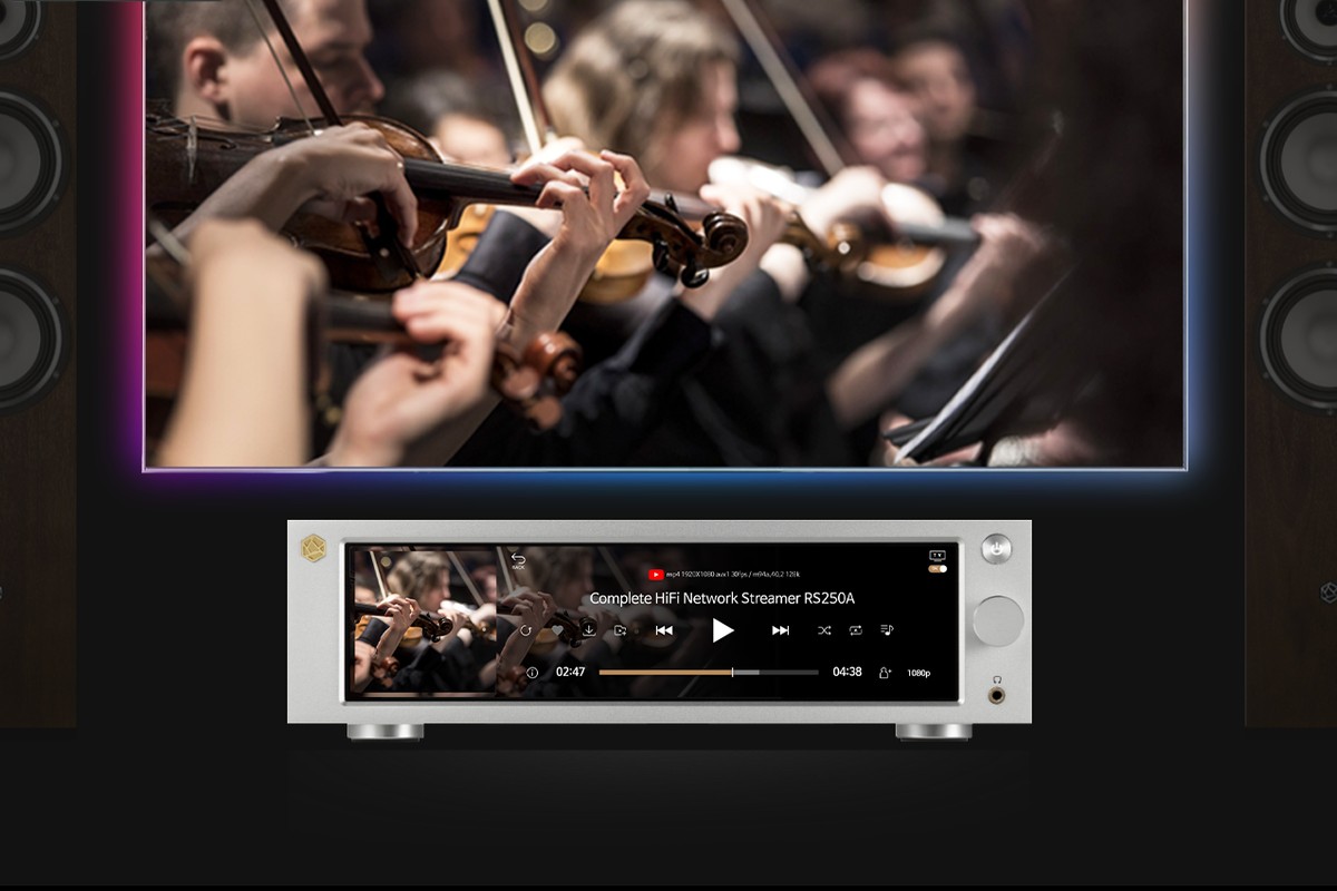 Streamer argento sotto un televisore che trasmette un concerto d'orchestra, tra due diffusori da pavimento