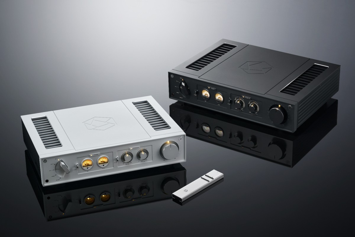 Two amplifiers, one silver and one black, with their remote control on a dark surface
