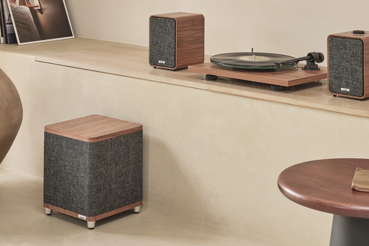 The RS1 subwoofer in walnut standing on the floor beneath a console, with two MR1 speakers and a turntable above