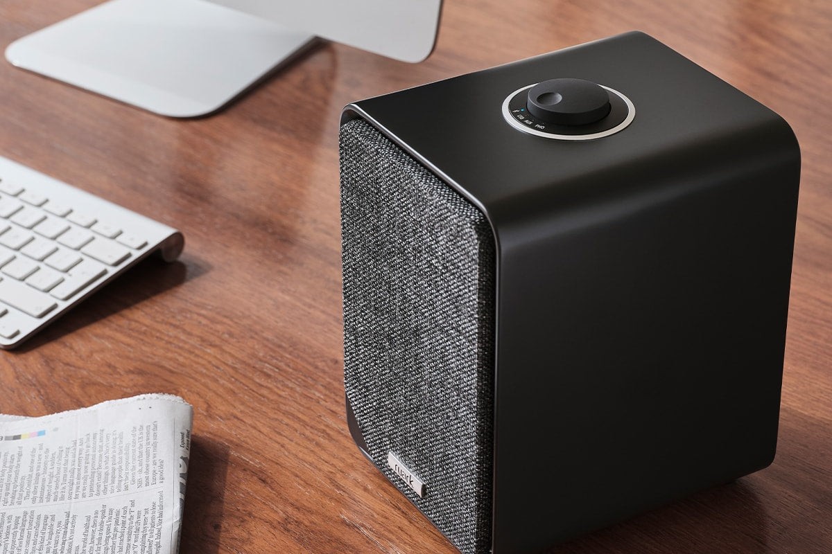 Primo piano di un diffusore attivo in finitura Satin Charcoal appoggiato su una scrivania, tastiera e schermo sullo sfondo