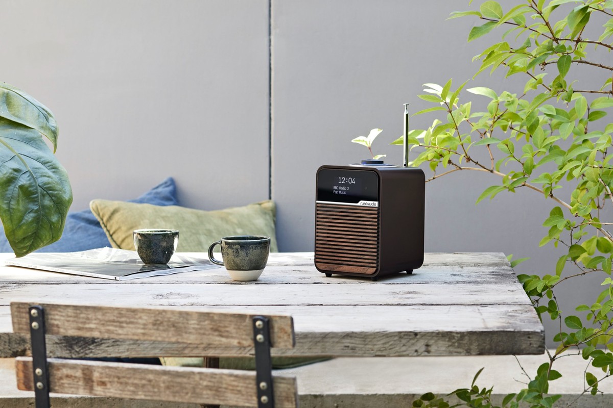 Radio posée sur une table de jardin en bois, antenne télescopique sortie, tasses et plantes autour
