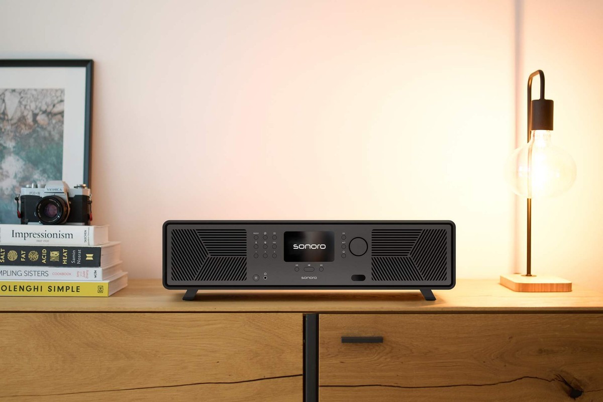 Système audio noir mat posé sur un buffet en chêne entre une pile de livres et une lampe à ampoule