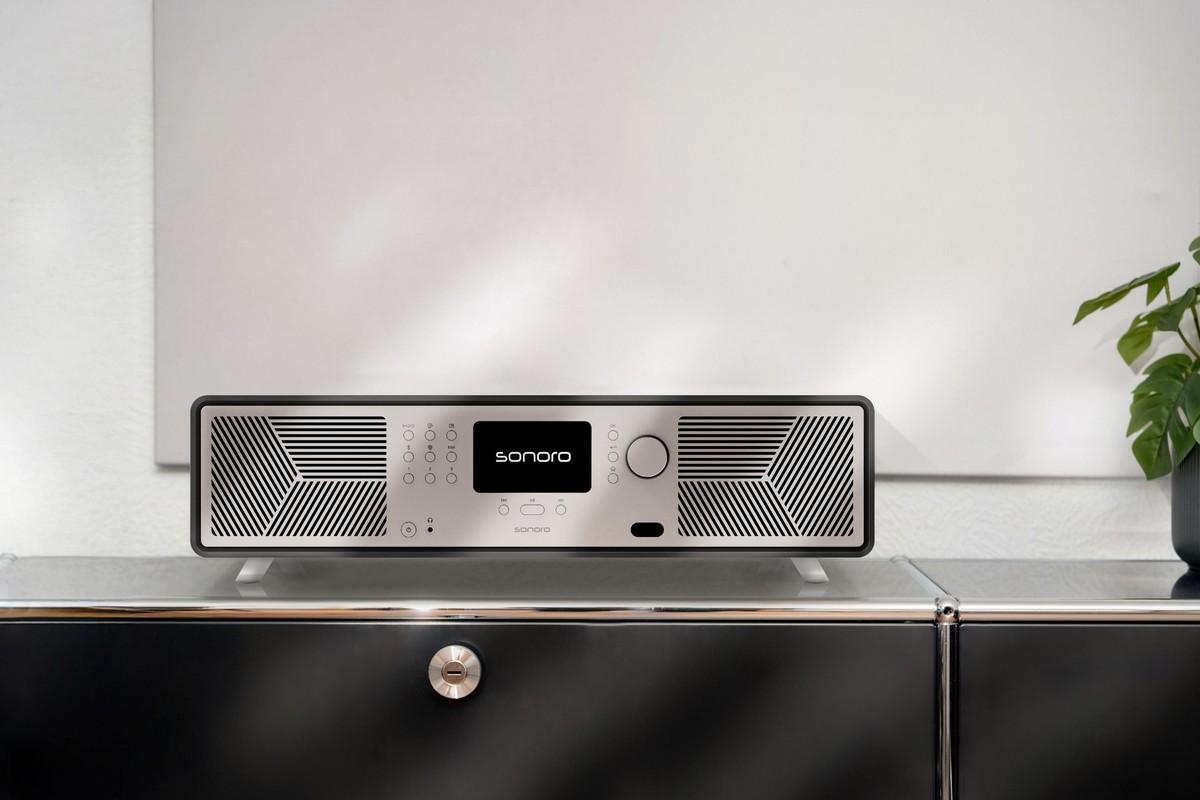 Système audio graphite à façade argent posé sur un meuble bas noir laqué devant un mur blanc