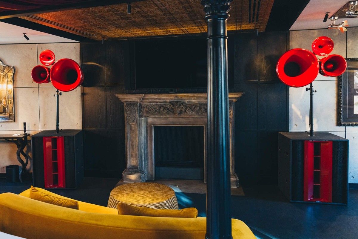 Deux caissons de basses noirs à pavillon rouge sous des enceintes à pavillons rouges dans un salon sombre avec cheminée