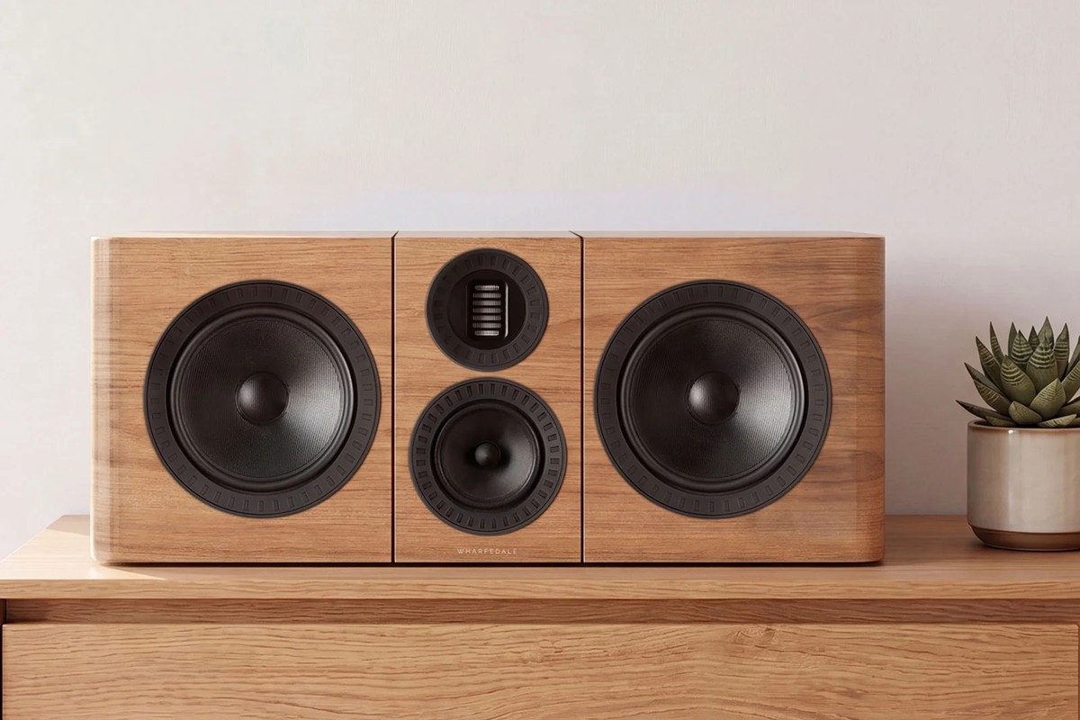 A light walnut centre speaker on a wooden cabinet, in front of a light wall and a plant