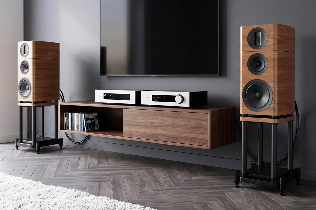 Two speakers on stands on either side of a television, with two amplifiers on a low cabinet