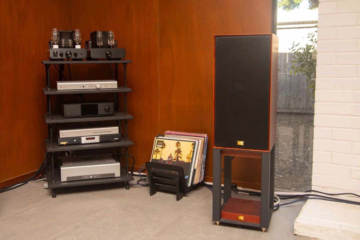 Speaker on a black stand next to a vinyl record crate and an electronics rack