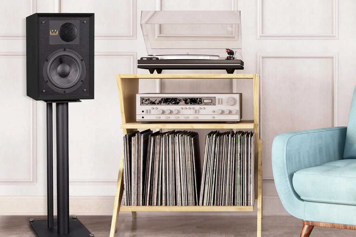 Black speaker on a stand in front of a light wall, next to an open cabinet with a turntable, amplifier and records