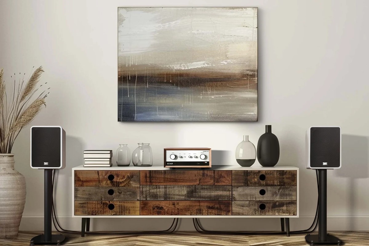 Two white stand-mounted speakers either side of a wooden sideboard, with an amplifier and vases