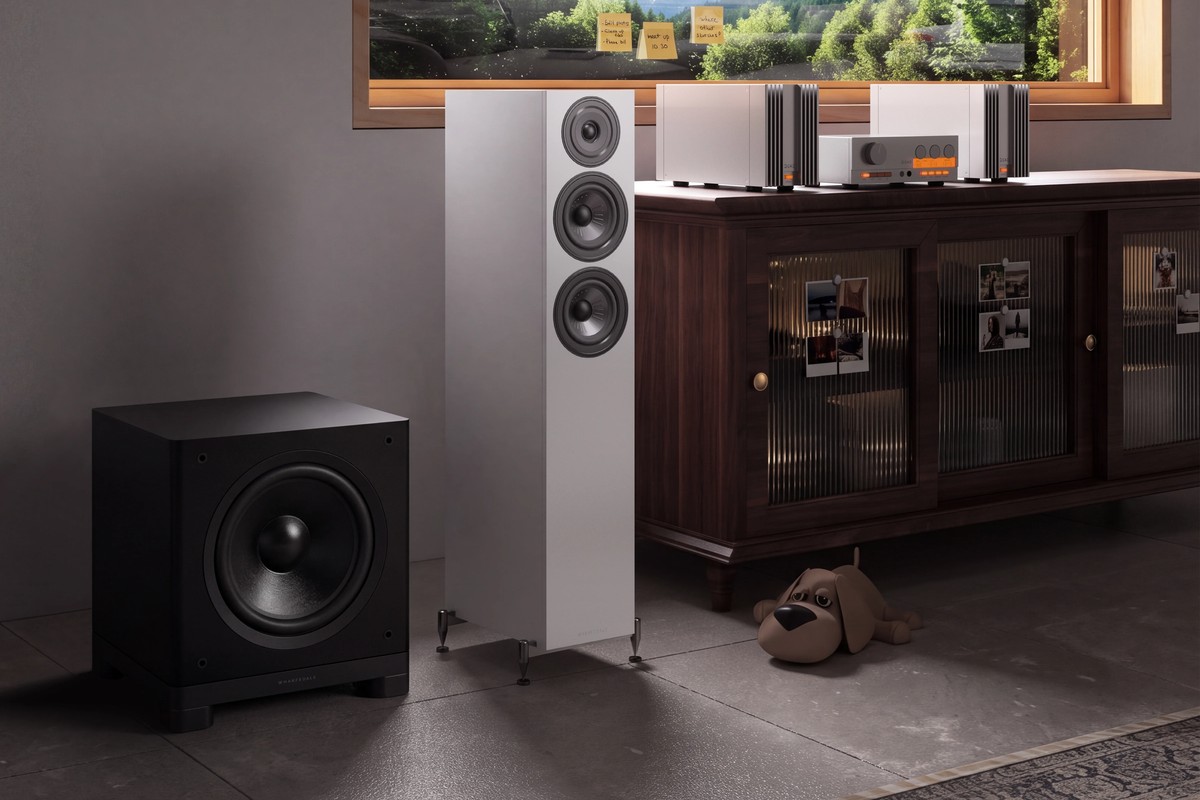 Black subwoofer on the floor next to a white floorstanding speaker and a wooden cabinet holding electronics