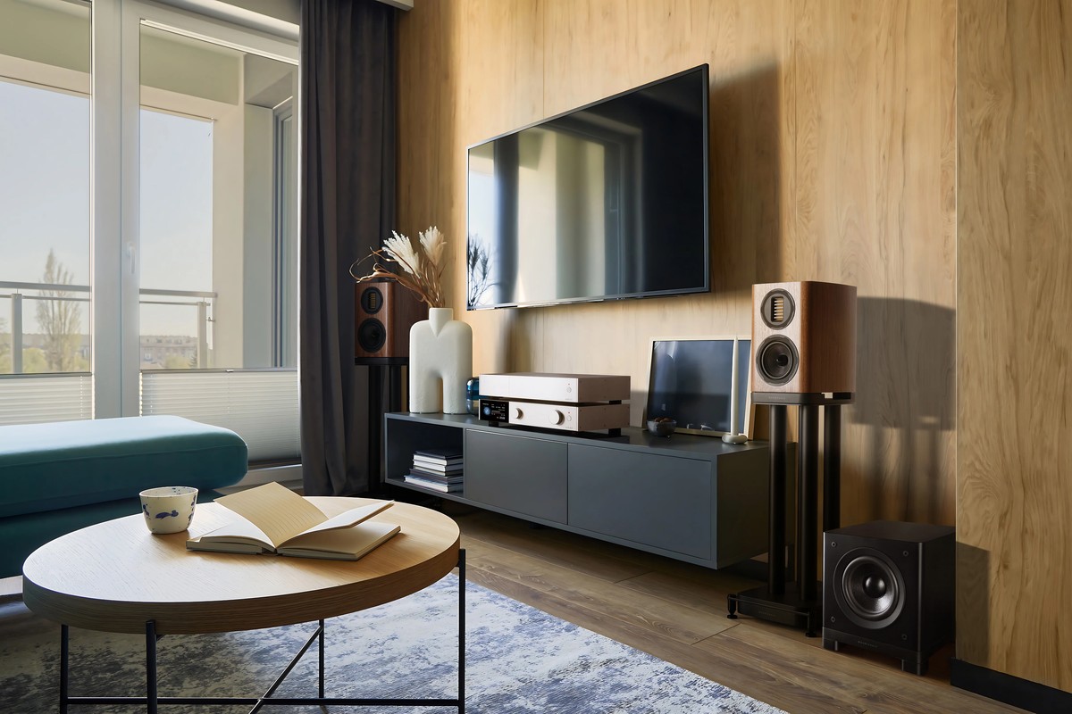 Living room with a wall-mounted TV, two compact speakers on stands, an amplifier and the black subwoofer on the floor