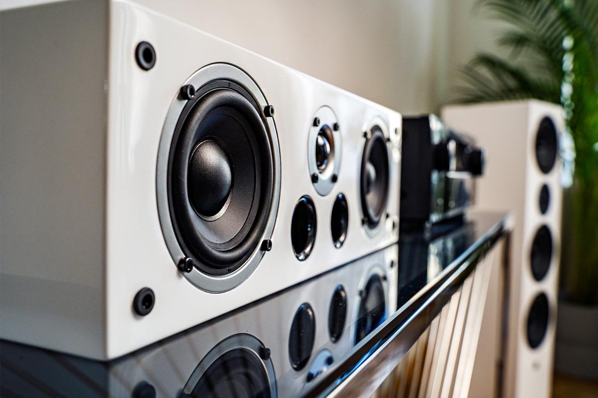 White lacquered centre speaker on a cabinet, in front of an amplifier and a floorstanding speaker