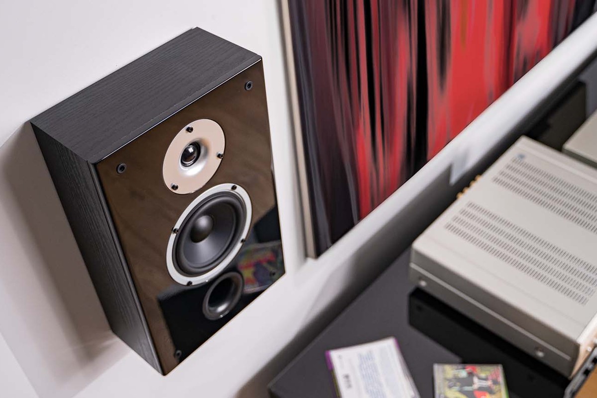 Black on-wall speaker mounted on the wall, seen at an angle, next to a hi-fi cabinet with a silver amplifier