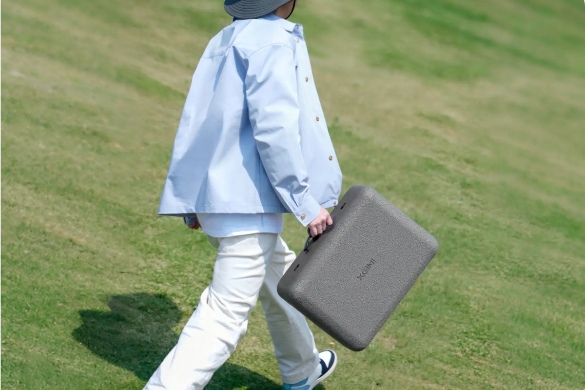Person in a hat walking across a lawn, grey carrying case in hand