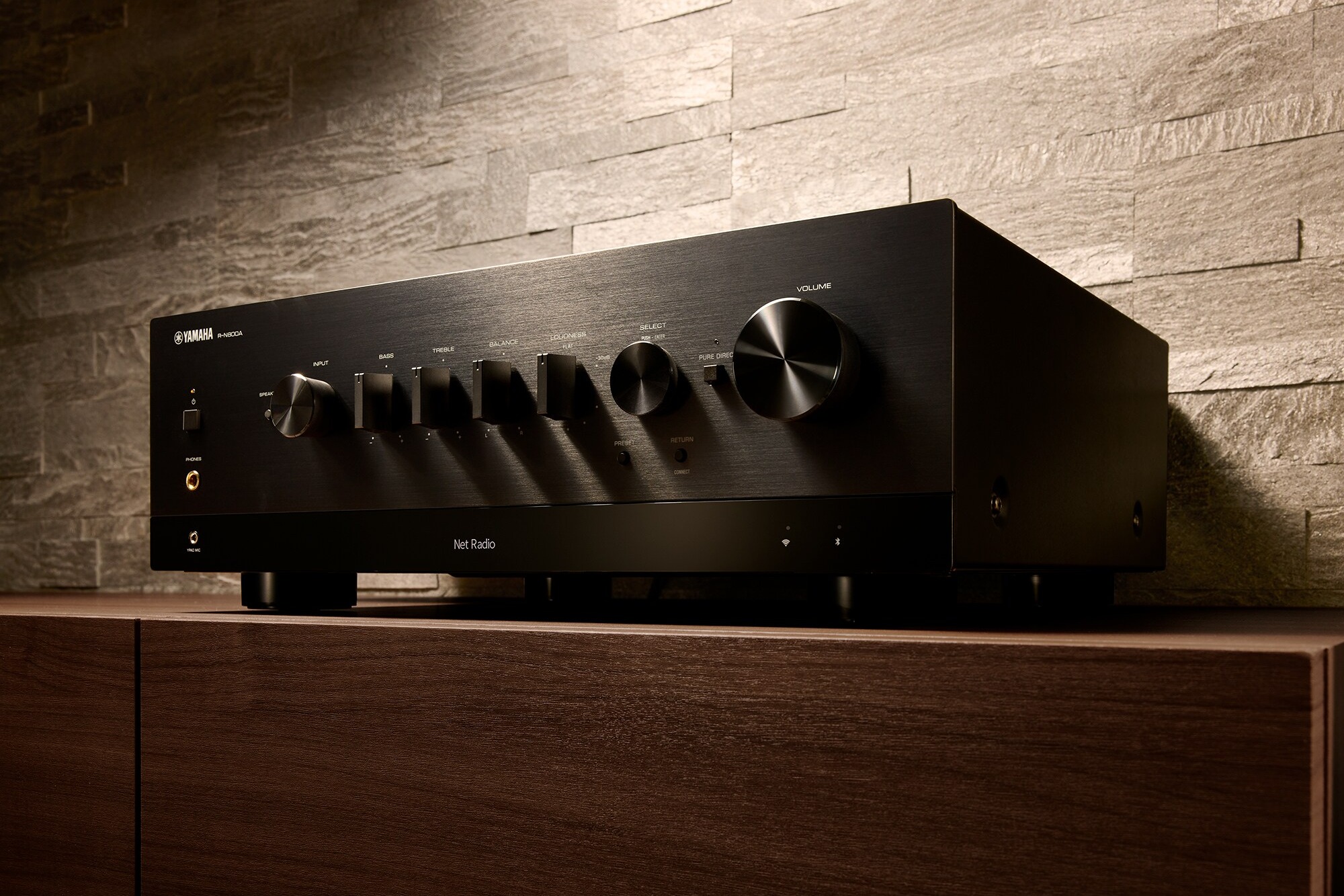 Ampli-tuner réseau noir posé sur un meuble en bois, mur de pierre en fond