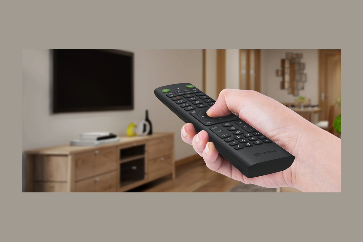 Main tenant une télécommande noire devant un meuble TV en bois et un téléviseur mural